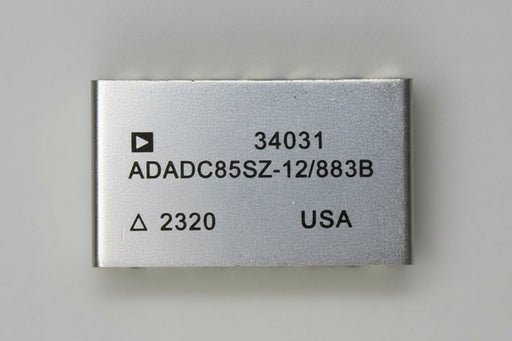 A rectangular silver metal electronic component with black printed text including '34031', 'AD2700UD', '/883B', '2320', and 'USA', photographed on a beige surface.