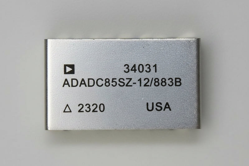 A rectangular silver metal electronic component with black printed text including '34031', 'AD2700UD', '/883B', '2320', and 'USA', photographed on a beige surface.