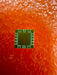 A small square electronic chip with a green and gold surface, placed on a textured orange-red background.