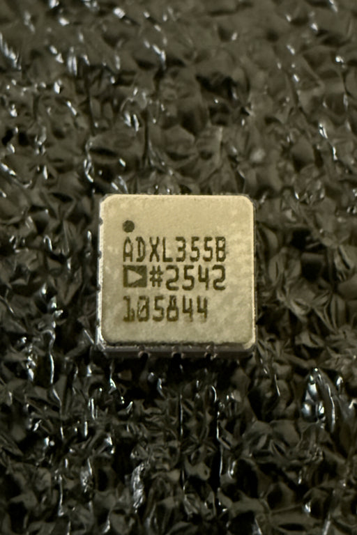 A square integrated circuit chip with black printed text reading 'ADXL355B #2542 105844' on a beige surface, set against a dark textured background.