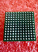 A square green electronic component with a grid of silver pins, photographed on a textured red background.