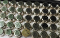 A close-up of many small cylindrical metal electronic components arranged in rows on a white surface. Most are silver with black printed text, and one near the bottom center is gold-toned.