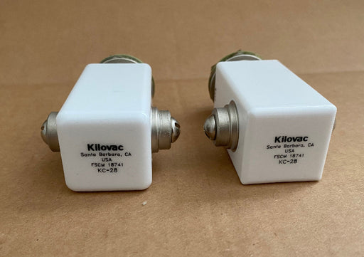 Two white Kilovac RF isolators with silver metal connectors, placed on a brown cardboard background.